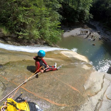 Person sitzt am Rand eines Wasserfalles und blickt auf einen Fluss in einer bewaldeten Umgebung.